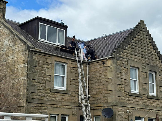 Two workers on a sloped stone roof use a ladder to doing roof maintenance