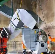 Workers in safety gear using cranes to install large industrial equipment with metal parts suspended above