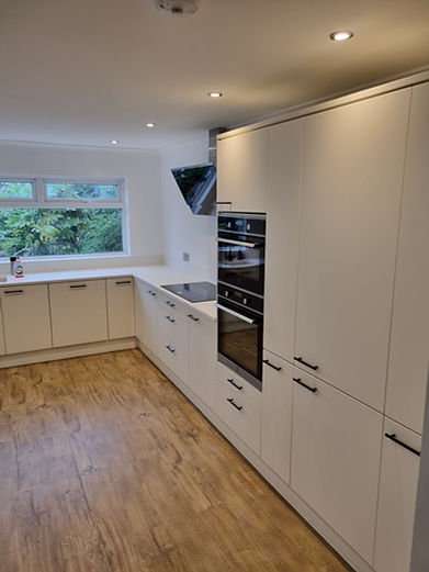 Modern kitchen featuring sleek white handleless cabinets, integrated double oven, electric hob, wooden flooring, and large window.