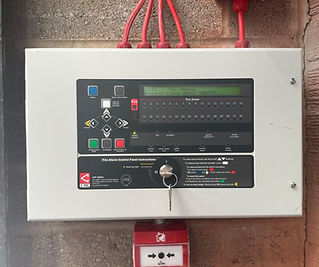 A fire alarm control panel with a digital display and various buttons, mounted on a wall