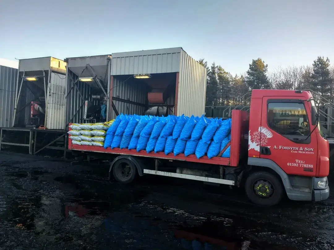 A red delivery truck loaded with yellow and blue sacks parked outside a coal facility