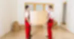 Two movers in red uniforms carrying a wooden dresser in a room