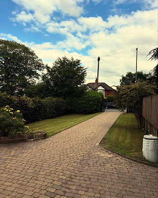 A paved driveway leads to a brick house, surrounded by lush greenery and trees