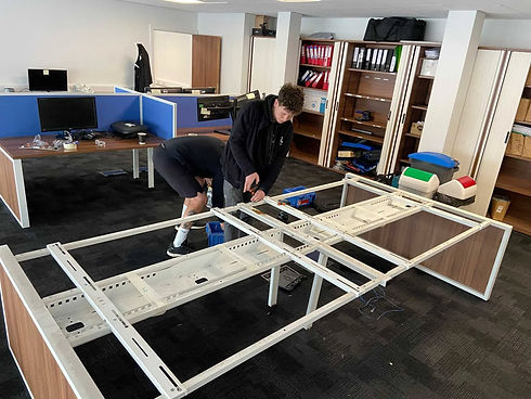 Two men assemble a white metal desk frame in a modern office with blue cubicles and shelves of binders