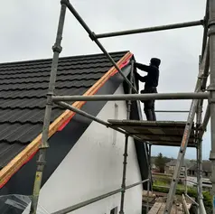 Roofer on scaffolding installing black wave-shaped tiles on the gable end of a white house