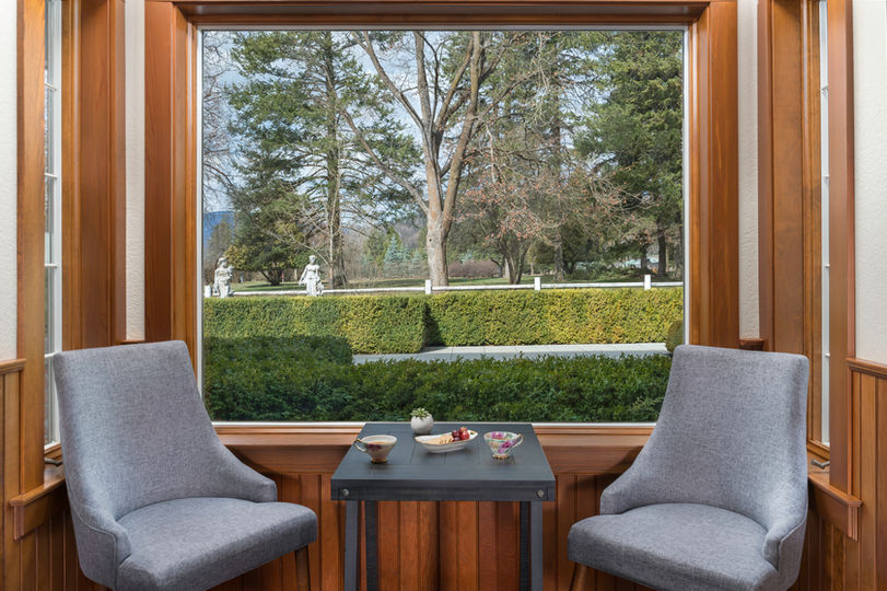 Comfortable sitting area with two gray chairs and a small table overlooking a garden and statues through a large window.
