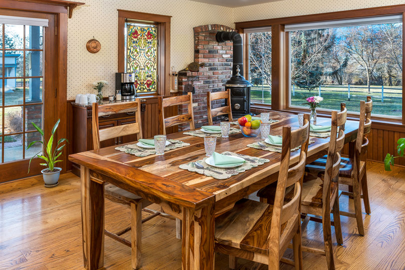 Rustic wooden dining table set for breakfast in a cozy room with large windows, a wood stove, and a stained glass window.