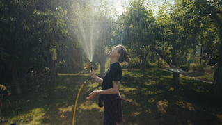 Woman looking up into hose water spray in sunny garden.