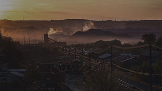Silhouetted industrial facility emits smoke at sunset, hazy mountains. Dystopia Feature Film.