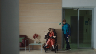 Woman in wheelchair talks with man in modern hallway near "RO" sign.