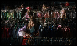 Young women browse clothing racks in a thrift store; one wears a feathered headdress — a still from 