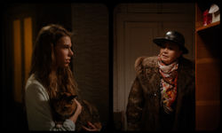 A young woman holding a cat speaks to an older woman in a fur coat and hat at a doorway — a still fr