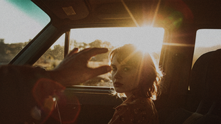 Young woman in car looks back, hand pinching sun. Dystopia Feature Film.