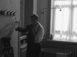 Man putting on jacket in dark, sparse room with bathtub.