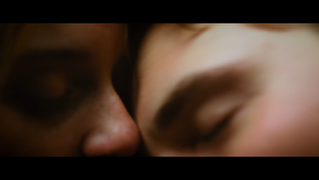 Two individuals' faces, eyes closed, intimately close in dark setting.