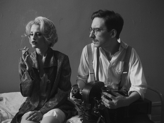 Blonde woman smoking cigarette, man holding camera, vintage black and white.