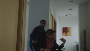 Man pushes surprised elderly woman in wheelchair along a light hallway.