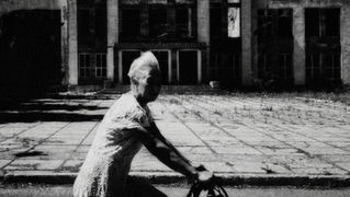 Elderly person on bicycle passing a dilapidated building