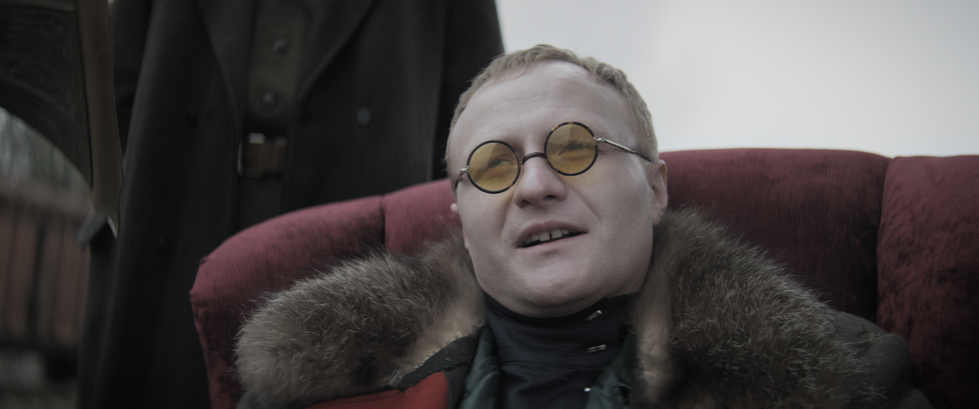 Блондин у жовтих окулярах, хутряному пальто, червоному кріслі. Історичний військовий фільм.