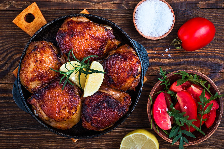 Roasted chicken in a skillet with lemon and rosemary. Side of sliced tomatoes and arugula in a bowl. Salt and tomato on wooden surface.