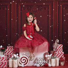 Smiling girl in a red holiday dress sitting among Christmas presents, candy canes, and festive decorations.