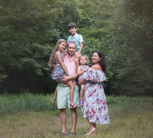 Family of five posing together in a wooded field during an outdoor portrait session, dressed in soft summer colors.