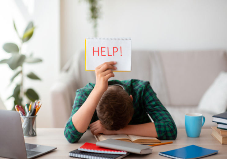 Person slumped over a desk with books, holding a sign reading "HELP!" in a cozy room. Meditation is the best self-help.