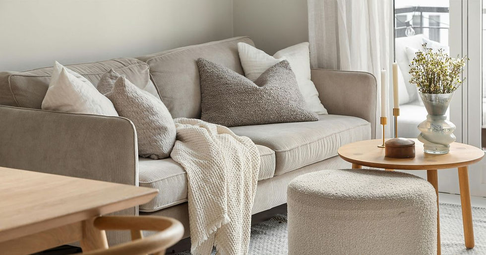 Cozy beige living room with corduroy sofa, textured pillows, knit throw, round table with vase and candles by bright window.
