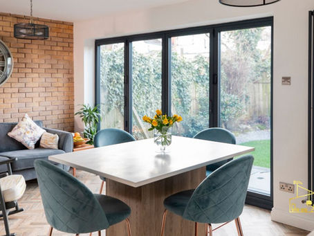 Bright dining room with teal chairs, gray sofa, brick wall clock, flower vase, and sliding doors to garden; EPIC watermark