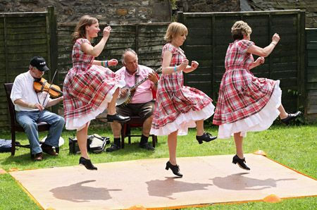 Three women in plaid dresses dance on a mat, while two men play violin and banjo. Outdoors on grass with a fenced background. Energetic mood.