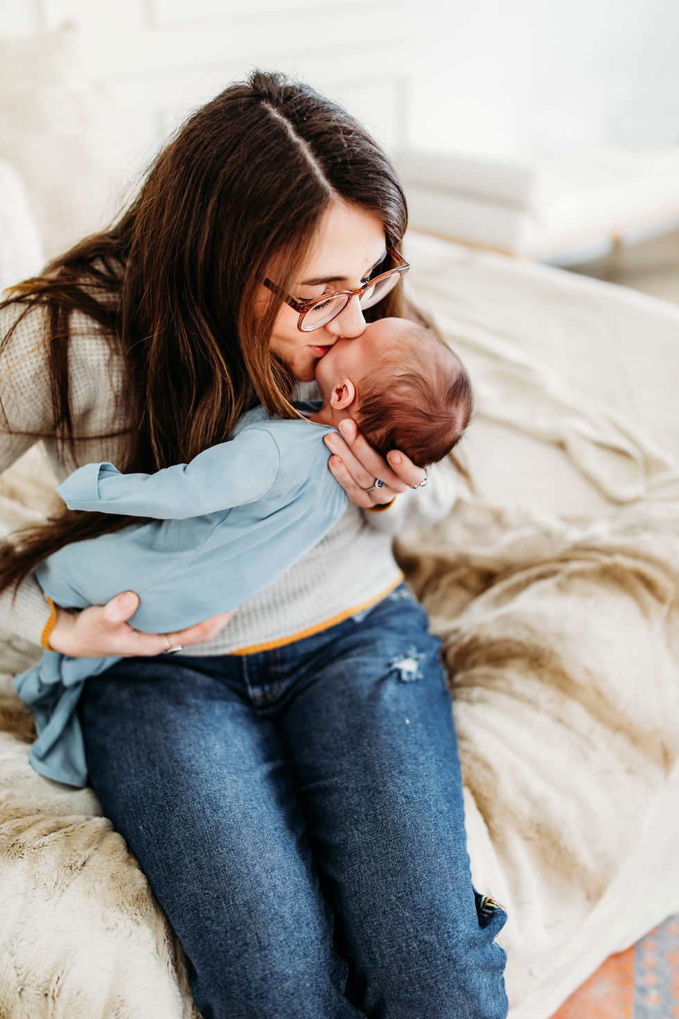 Woman lovingly kisses a baby wearing blue, seated on a beige couch. The setting is cozy and bright, with a warm, affectionate mood.