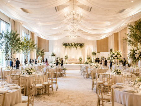 Elegant wedding reception hall with soft white drapes, floral centerpieces, crystal chandeliers, and a beautifully designed stage creating a warm and welcoming atmosphere.