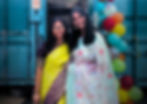 Two women in vibrant sarees smile in front of a teal backdrop with colorful balloons. A sign reads "Where Burrito Meets Bollywood" at Salsa Samosa in Bristol