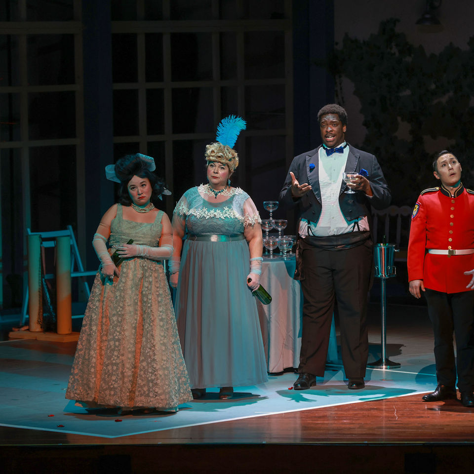 Opera performers in costume on stage at a formal dinner scene.