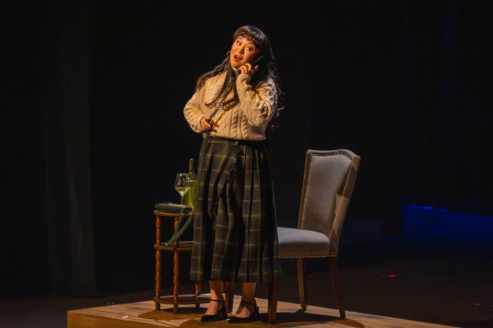 Woman singing on stage next to a chair and small table.