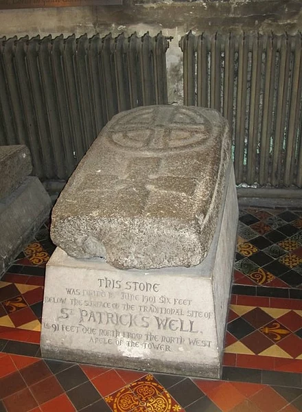 A large, ancient stone slab with a carved cross on its surface rests on a pedestal inside a historical building. The pedestal bears an inscription stating that the stone was found in June 1901, six feet below the surface at the traditional site of St. Patrick's Well, located in St. Patrick's Cathedral, Dublin, Ireland. The floor around the pedestal is decorated with colorful geometric tiles, and a radiator is visible in the background.