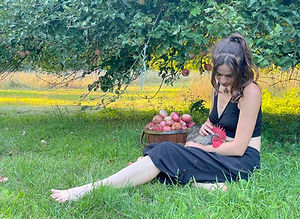 A young brunette woman sits on the ground under an apple tree next to a bucket of apples while she looks down sweetly at the rooster she is holding.