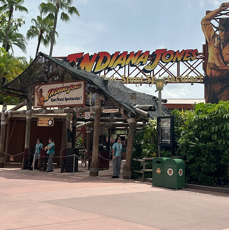 Adventure-themed entrance with “Indiana Jones Epic Stunt Spectacular!” sign featuring rugged typography, palm trees, and bamboo structures evoking an action movie set.