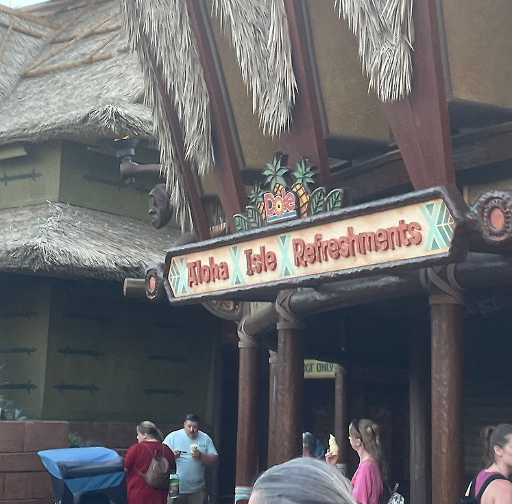 Aloha Isle in Adventureland, a tropical-themed kiosk with a tiki-style roof, wooden details, and guests enjoying Dole Whip treats.