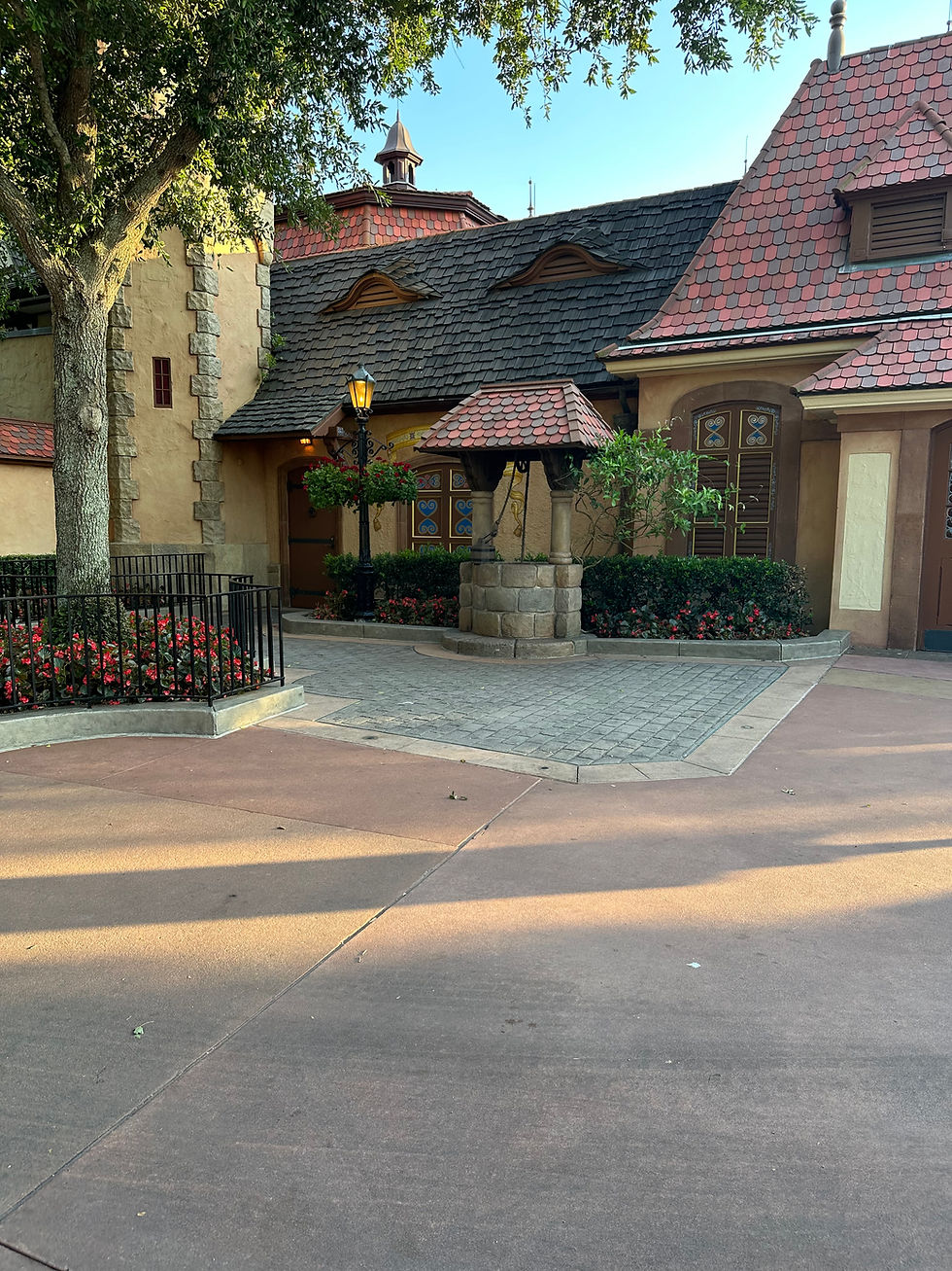 Wishing well with German Style buildings in the background and a tree, flowers and black iron fence to the side