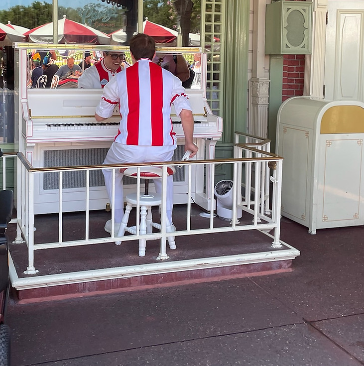 Street pianist performing at Casey’s Corner in Magic Kingdom, seated at a red-and-white vintage piano outside the baseball-themed eatery, entertaining guests with lively tunes.