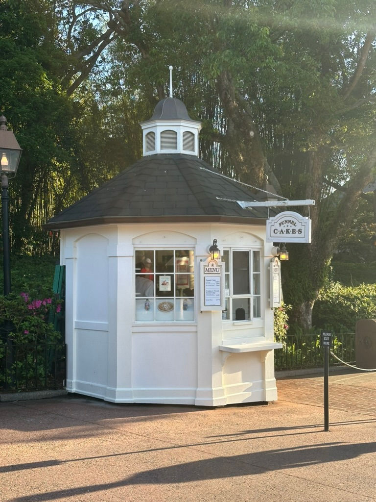 Funnel Cake stand at EPCOT, featuring a charming colonial-style kiosk with a decorative sign and brick details, serving freshly made funnel cakes topped with sweet treats in the American Adventure Pavilion.