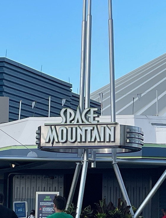 Entrance to Space Mountain at Magic Kingdom with the iconic futuristic sign, sleek white spires of the ride structure in the background, and guests lining up beneath the covered queue area.