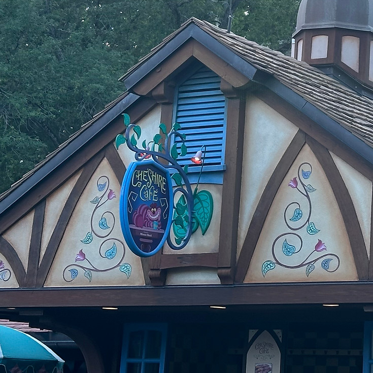 Cheshire Cat sign near the Mirabel meet and greet at Magic Kingdom, with a mischievous grin and vibrant colors nestled in the whimsical foliage of Fantasyland.