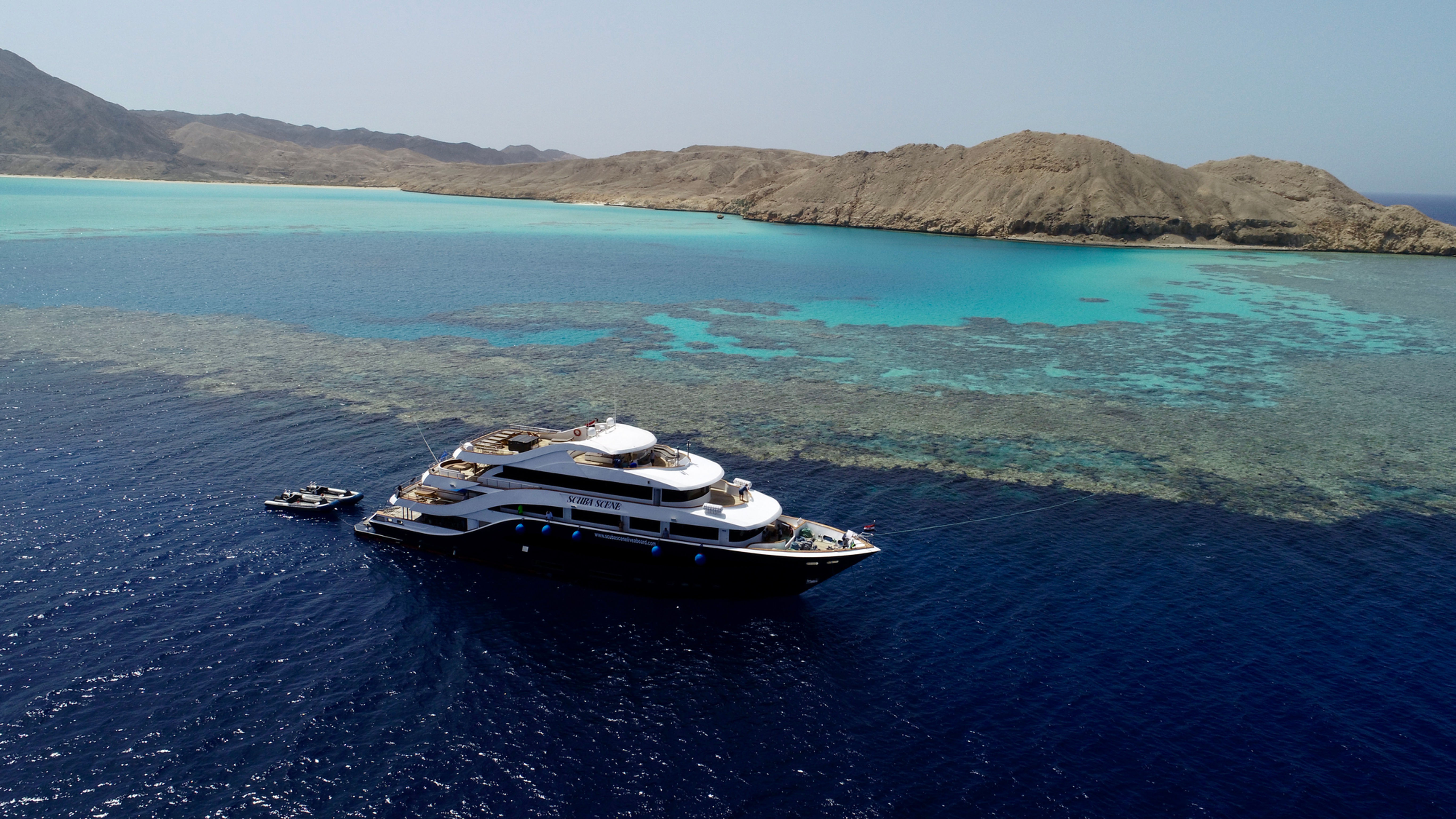 Luxury yacht cruising turquoise waters near a coral reef and rocky island.
