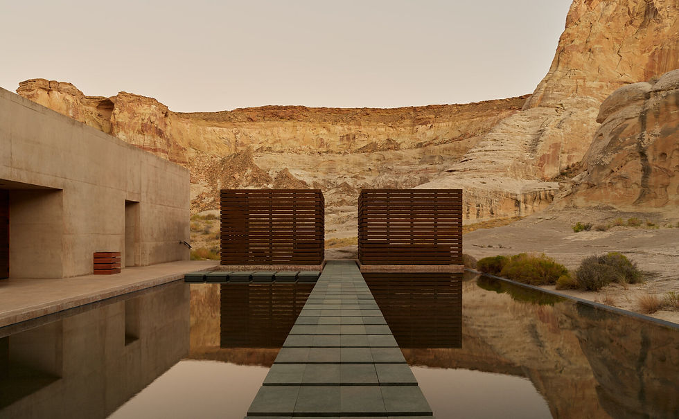 Stone pathway leads over a reflective pond between modern wooden structures. Desert cliffs in the background under a soft, golden sky.