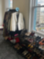 Clothing rack with a white blazer and assorted garments. Shoe rack with various shoes. Bright room with large windows showing urban view.