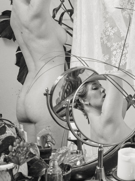 a black and white image taken looking into multiple mirrors with a nude woman putting her hair up and looking over her shoulder