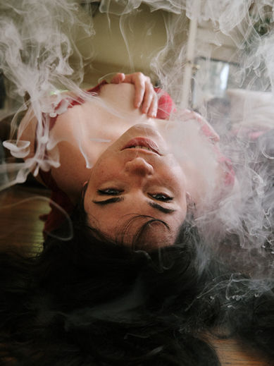 a woman with black hair and blue eyes lying on the floor upside and arching her back while looking up into the camera with smoke in the air around her