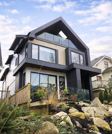 Northeast angled view of the duplex showing the gable roof massing, layered balconies, and glass guardrail at the sheltered attic deck above the sloped boulder landscaping.